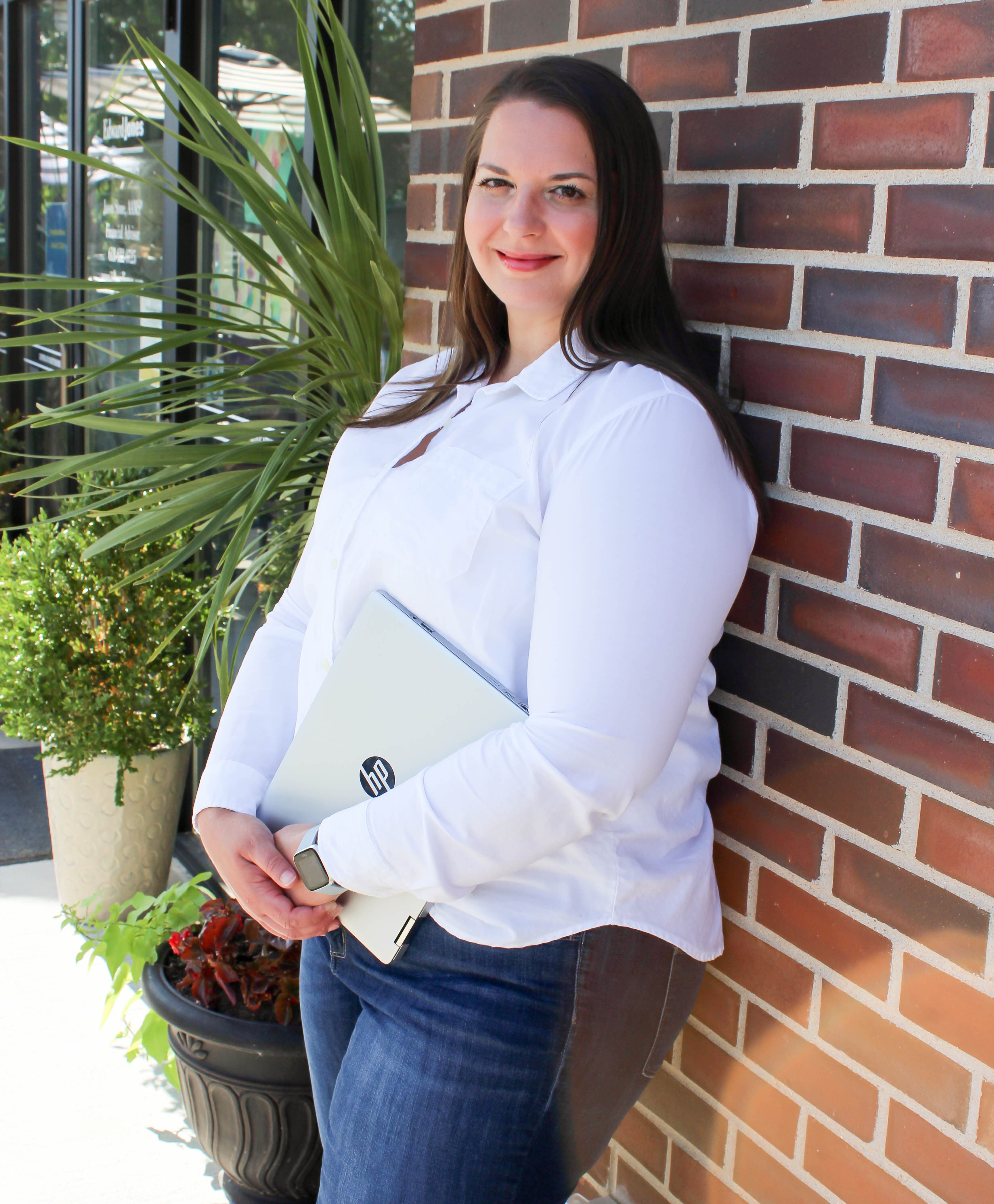 Smiling woman in white shirt leans against a brick wall holding a silver laptop.